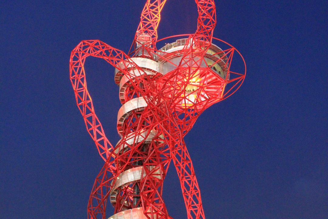 The red steel lattice of the ArcelorMittal Orbit lit up against a dark blue night sky, with its spiral walkway wrapped round the tower.