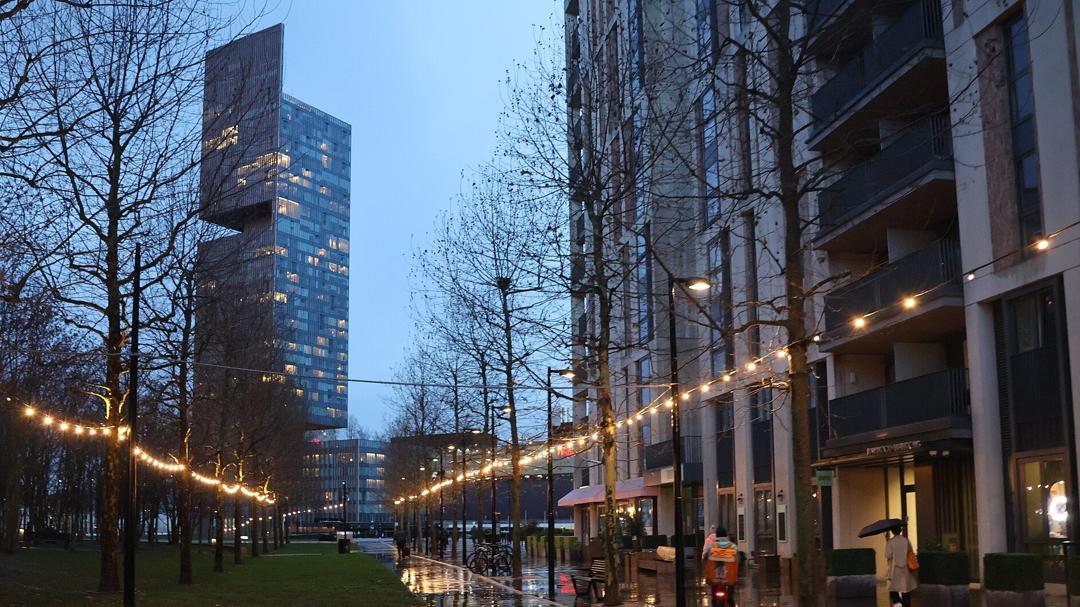 Festoon lights along the edge of Victory Park at dusk, with shopfronts on the right and Manhattan Loft Gardens behind
