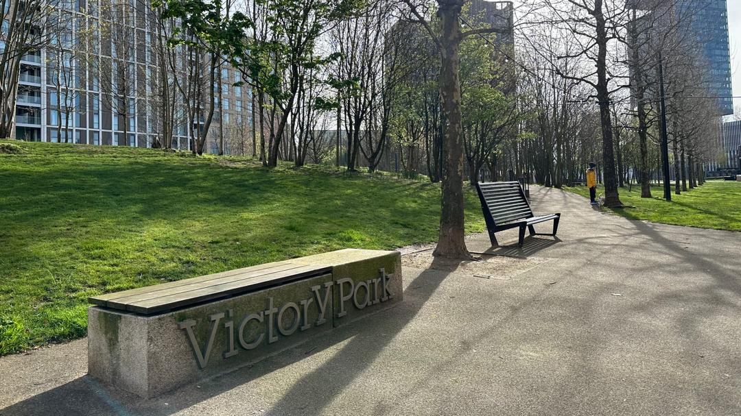 The Victory Park sign on a stone bench at the park's entrance in East Village, with trees and the estate's towers behind